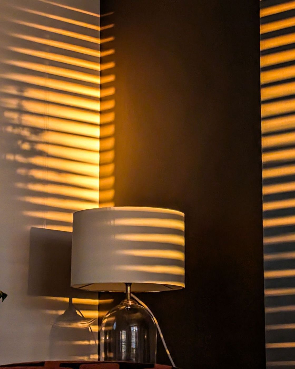LUMI Ambient Smart Lamp: a white drum-shade lamp on a glass base, glowing in a dark room striped with window light